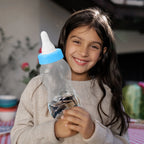 Child holding a bottle piggy-bank money box coin bank with a blue cap, smiling outdoors.