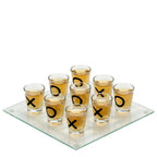 A drinking tic tac toe game with a glass board and multiple shot glasses arranged on top, with the shot glasses having 'X' and 'O' marks.